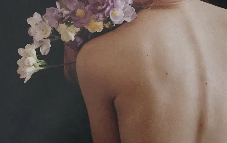Mujer de espaldas con cabello rosa adornado con flores, fondo oscuro que resalta la estética natural y delicada de la imagen.