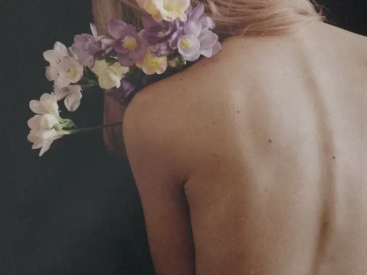 Mujer de espaldas con cabello rosa adornado con flores, fondo oscuro que resalta la estética natural y delicada de la imagen.